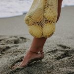 Barefoot stepping on sandy beach with lemons in mesh bag, evoking summer vibes.