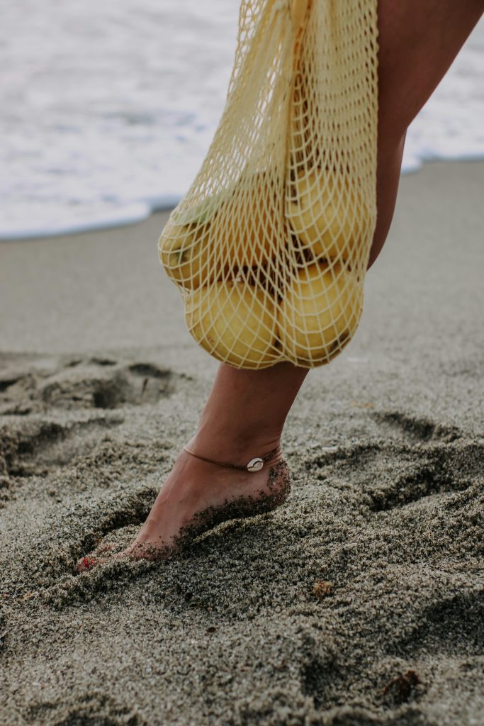 Barefoot stepping on sandy beach with lemons in mesh bag, evoking summer vibes.